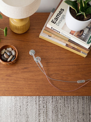 A pair of translucent light gray wired earbuds with a matching inline remote and cable, laid out on a warm brown wooden tabletop. The scene is styled with a yellow table lamp, a small bowl of pebbles, a potted plant, and a stack of Eames Design books.