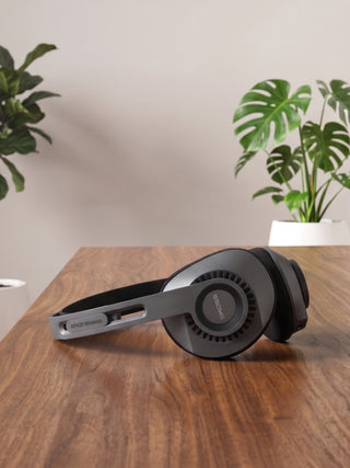 A pair of over-ear headphones resting on a wooden table, with potted green plants in the background against a neutral wall.