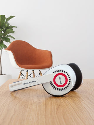 White and black KPVDS wireless headphones with a red circular logo, resting on a light wood table in front of a modern orange and white chair.