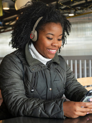 A woman with curly black hair wearing gray over-ear headphones and a dark quilted jacket sits at a table, smiling while looking at a smartphone in a cafe setting.