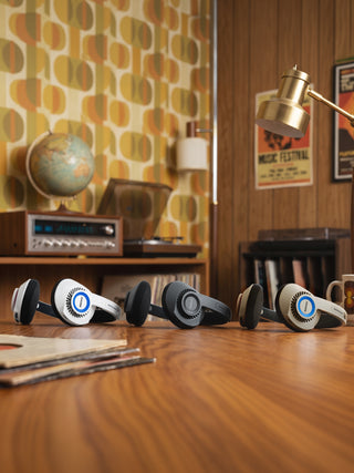 Four vintage-style over-ear headphones in white, black, and gray are lined up on a wooden table in a retro-themed room with patterned wallpaper, a record player, and a desk lamp.