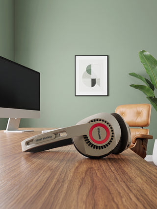 A pair of Sennheiser wireless over-ear headphones resting on a wooden desk in a modern home office setting with a computer monitor, chair, and framed wall art.