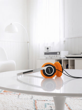 Orange and silver over-ear headphones with black ear cushions resting on a white table in a bright, modern room.