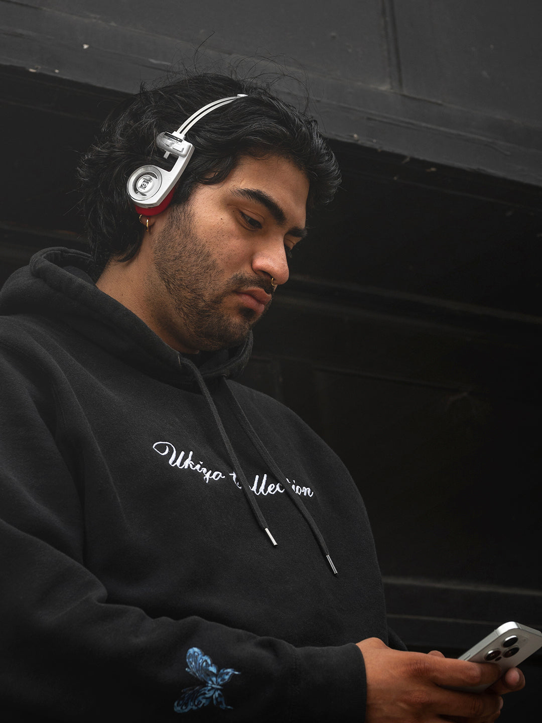 Man wearing headphones and a black hoodie with text, looking at a phone.