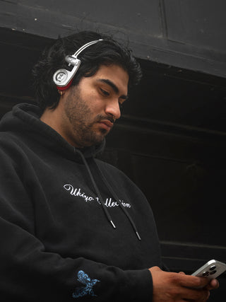 Man wearing headphones and a black hoodie with text, looking at a phone.