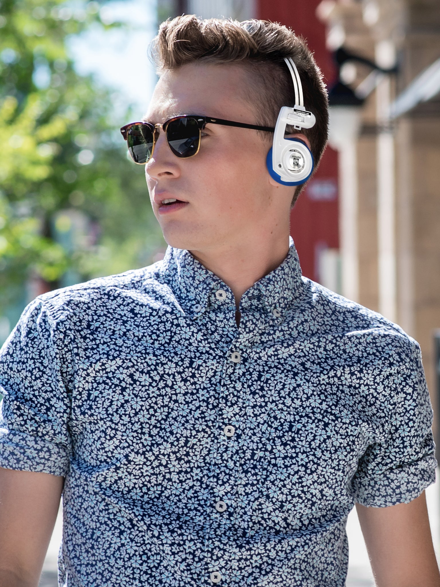 Man wearing sunglasses and a patterned shirt with headphones around his neck outdoors.