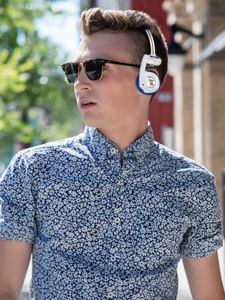 Man wearing sunglasses and a patterned shirt with headphones around his neck outdoors.